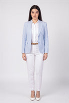 A woman modeling a light blue linen blazer over a white shirt and white trousers, styled with a thin brown belt and white heels, against a plain light gray studio background.