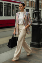 A woman walking on a city street wearing a striped linen blazer with pastel pink, blue, orange, and green stripes over a white top and cream wide-leg trousers, carrying a black tote bag.