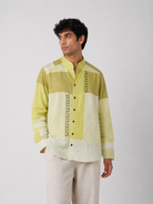 A man wearing a light green and off-white patchwork long-sleeve shirt with green leaf embroidery down the front, paired with off-white trousers, against a plain light gray background.