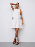 A woman modeling a sleeveless white linen dress with embroidered daisy motifs on the front, paired with black strappy sandals, set against a plain light gray wall.