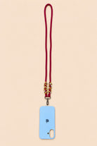 A light blue phone case with a matching red cord lanyard and gold-toned decorative beads, shown on a solid pale peach background.